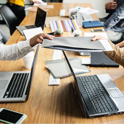 People sat at a desk with laptops, a folder is being passed from someone on the right hand side of the image to a person on the left. 