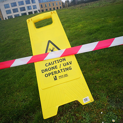 A yellow caution drone / UAV operating warning sign on a grass field with a building in the background. 
