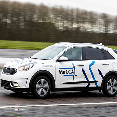 A white car with the MuCCA logo on the side drives along a road. 