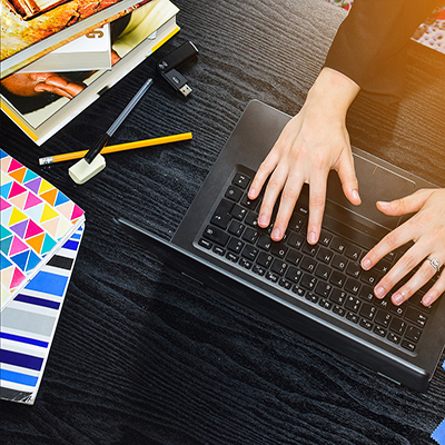 A woman types on a laptop. To her left there are colourful notebooks, pens and some study books. 